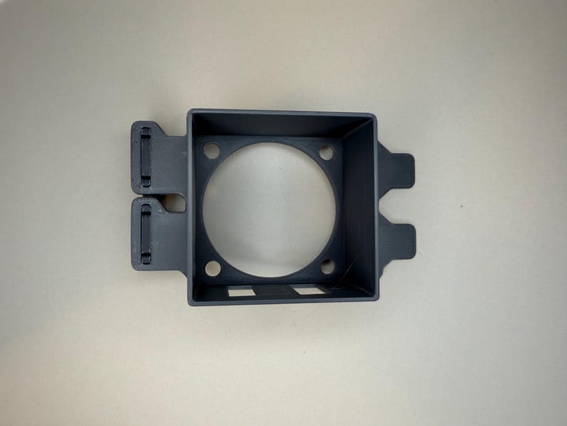 May include: A dark grey, rectangular plastic component with a circular aperture in the centre. It features two rectangular cutouts and several small holes. Two tabs extend from the side, against a light background.