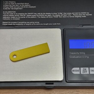 May include: A small, rectangular, yellow plastic tag with a single circular hole at one end, resting on a black digital scale with a silver weighing platform. The scale display shows a weight of 10.0 grams with a capacity of 500g.