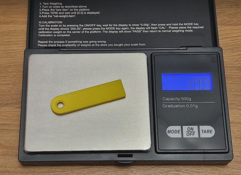 May include: A small, rectangular, yellow plastic tag with a single circular hole at one end, resting on a black digital scale with a silver weighing platform. The scale display shows a weight of 10.0 grams with a capacity of 500g.