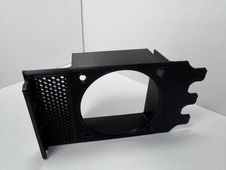 May include: A black 3D-printed bracket with a large circular opening and a perforated side panel. The bracket has several mounting holes and a unique, stylized edge design. The object is made of black plastic.
