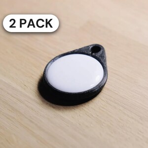 May include: A black teardrop-shaped tag with a white circular center and a small hole at the top for attachment. The tag features a textured finish and is labeled with the text 2 PACK in a white rounded rectangular box.