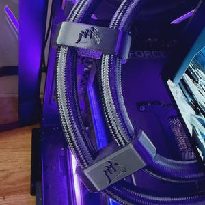 May include: A close-up of a computer's internal components, showing a liquid cooling system with braided tubing and black brackets. The Corsair logo is visible on the brackets. The background shows other computer parts and a screen.