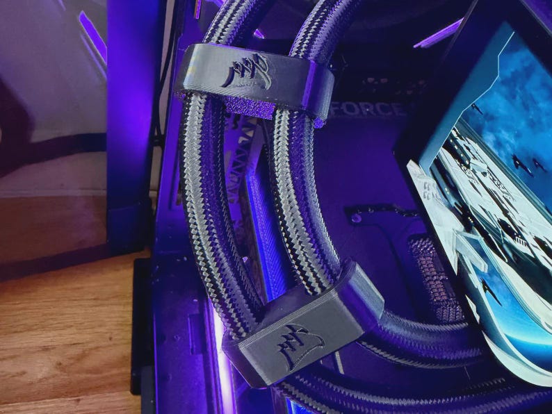May include: A close-up of a computer's internal components, showing a liquid cooling system with braided tubing and black brackets. The Corsair logo is visible on the brackets. The background shows other computer parts and a screen.