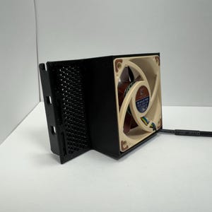 May include: A black computer fan with a beige and brown fan blade. The fan is housed in a black enclosure with a mesh side. A black cable extends from the fan.