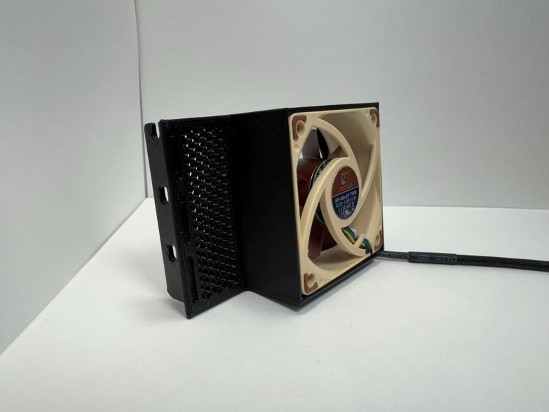 May include: A black computer fan with a beige and brown fan blade. The fan is housed in a black enclosure with a mesh side. A black cable extends from the fan.