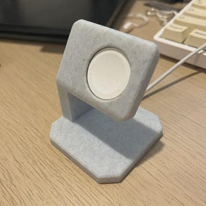 May include: A light grey, speckled Apple Watch charging stand. The stand has a square top with a circular indentation to hold the watch. The base is a geometric shape. A white watch is in the stand.