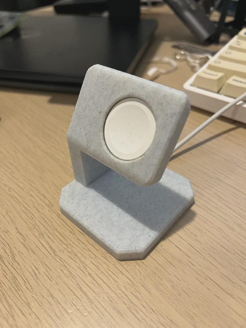 May include: A light grey, speckled Apple Watch charging stand. The stand has a square top with a circular indentation to hold the watch. The base is a geometric shape. A white watch is in the stand.