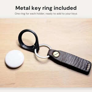 May include: A white circular tracking device next to a dark brown leather keychain with a brass rivet and a silver metal key ring. The text Metal key ring included and One ring for each holder, ready to add to your keys is visible.