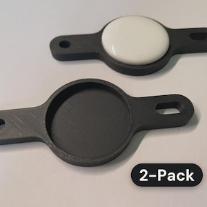 May include: A 2-pack of black plastic mounts, each featuring a central circular recessed area and two side tabs with holes for attachment. One mount contains a smooth, white, circular insert, while the other is empty.