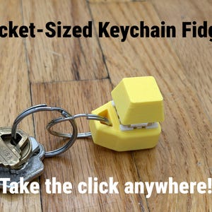 K&ouml;nnte beinhalten: Ein gelber, taschengro&szlig;er Schl&uuml;sselanh&auml;nger-Fidget mit einem Tastenkappendesign, befestigt an einem Schl&uuml;sselring mit Schl&uuml;sseln. Der Text "Pocket-Sized Keychain Fidget" und "Take the click anywhere!" sind sichtbar.