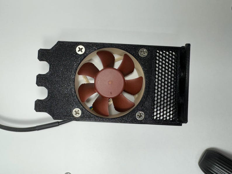 May include: A black computer fan assembly with a brown fan and a black mesh cover. The fan is secured with four screws. The assembly has a unique shape with a curved edge and a wire extending from it.