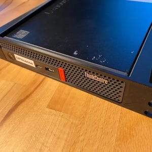 Könnte beinhalten: Ein schwarzer Lenovo ThinkCentre Computer in einem Rack-Gehäuse. Die Frontplatte verfügt über ein perforiertes Gitter, einen USB-Anschluss, einen kleinen roten Akzent und ein weißes Etikett mit dem Text OPNsense. Das ThinkCentre-Logo ist sichtbar.