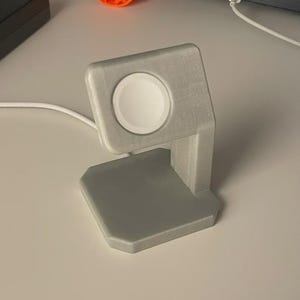 May include: A grey 3D-printed Apple Watch charging stand. The stand features a square base, a vertical support, and a circular charging area. A white charging cable is connected. An orange laptop stand is also visible.