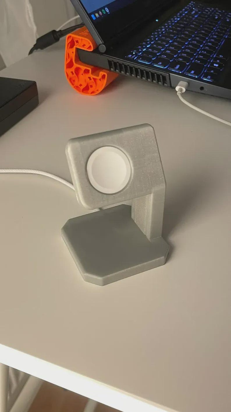 May include: A grey 3D-printed Apple Watch charging stand. The stand features a square base, a vertical support, and a circular charging area. A white charging cable is connected. An orange laptop stand is also visible.