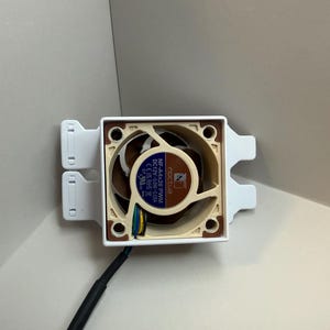 May include: A computer fan with a brown and beige fan blade design, mounted in a white square frame. The fan has a black cable and is designed for cooling computer components. The fan is labelled "Noctua".