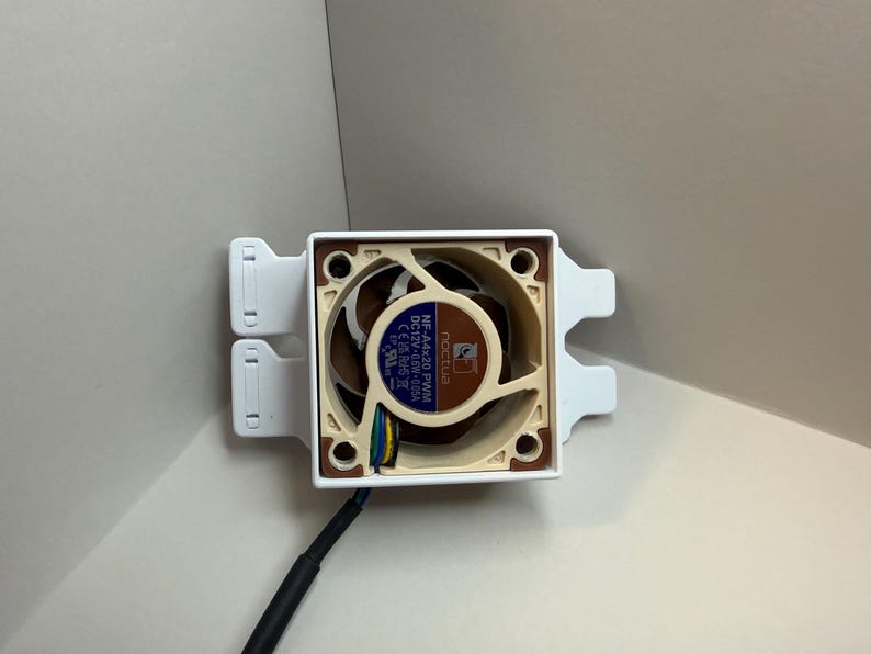 May include: A computer fan with a brown and beige fan blade design, mounted in a white square frame. The fan has a black cable and is designed for cooling computer components. The fan is labelled "Noctua".
