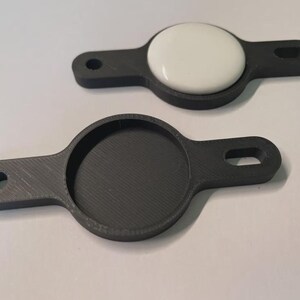 May include: Two dark grey 3D-printed holders for a round electronic device. One holder contains a white, circular device. Each holder has a circular recess and a handle with a hole for attachment. The holders are designed to secure and protect the device.