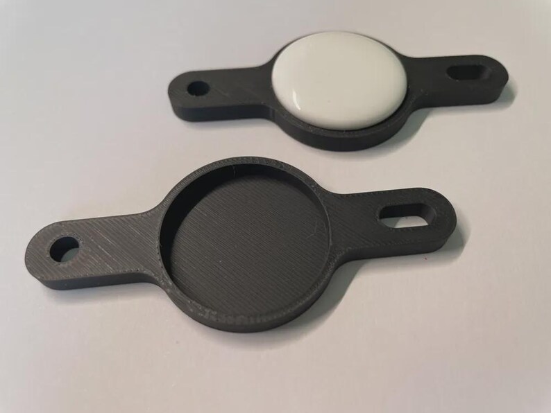May include: Two dark grey 3D-printed holders for a round electronic device. One holder contains a white, circular device. Each holder has a circular recess and a handle with a hole for attachment. The holders are designed to secure and protect the device.