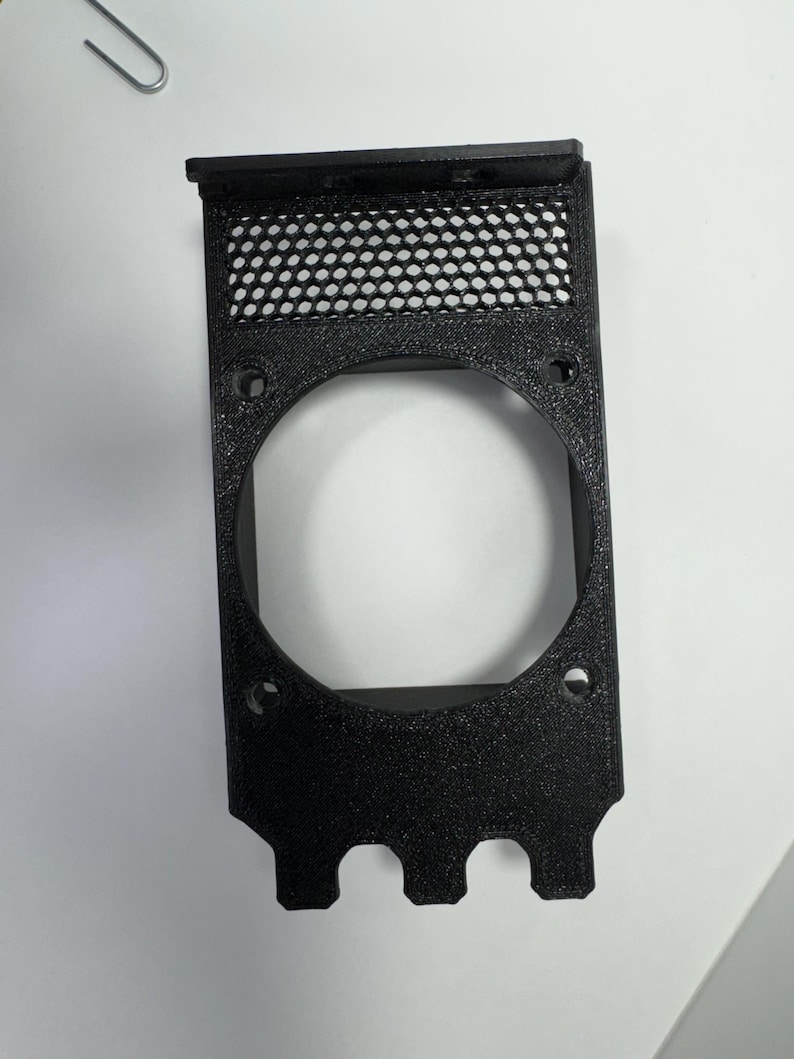 May include: A black, rectangular plastic component with a large circular opening and a mesh screen at the top. The component has several small holes and three rounded protrusions at the bottom.