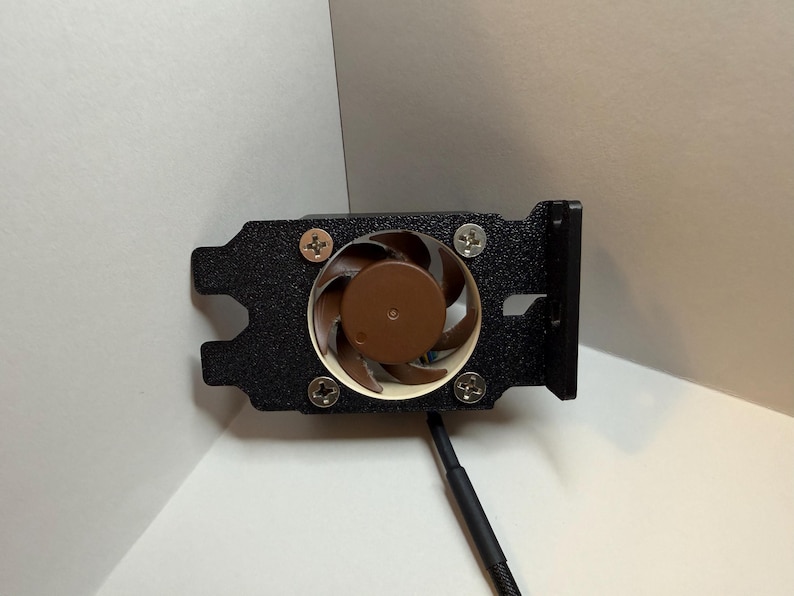 May include: A black computer fan with a brown central element and four silver screws. The fan is mounted on a black bracket with a cable extending from the bottom. Designed for computer cooling.