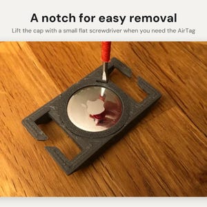 May include: A dark gray rectangular holder for an Apple AirTag with a central circular cutout. A small flathead screwdriver is inserted into a notch on the holder. Text reads: A notch for easy removal. Lift the cap with a small flat screwdriver when you need the AirTag.