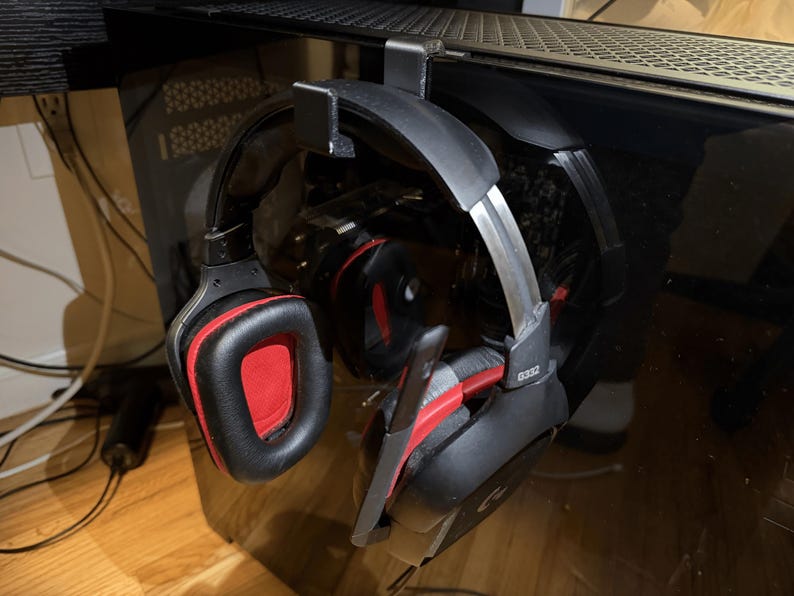 Op de afbeelding: Zwarte en rode gamingheadset die aan een zwarte metalen haak hangt. De headset heeft grote oorschelpen met rode vulling en een microfoon. De headset is ontworpen voor gaming en is bevestigd aan een computer.