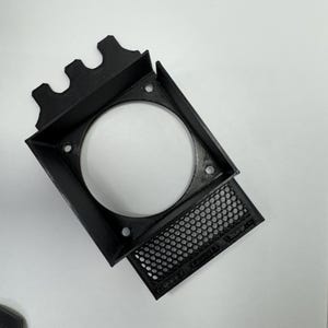 May include: A black, 3D-printed fan shroud with a square frame and a circular opening. The shroud has a perforated panel and mounting holes. The design includes a series of decorative peaks on one side.