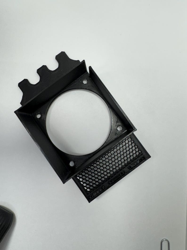 May include: A black, 3D-printed fan shroud with a square frame and a circular opening. The shroud has a perforated panel and mounting holes. The design includes a series of decorative peaks on one side.