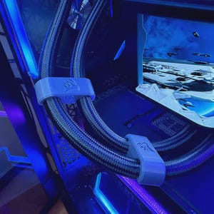 May include: A close-up of a computer's internal components, with a curved monitor showing a futuristic scene. Braided black and grey tubes are held by light grey clips with a logo. The scene is illuminated with blue LED lighting.