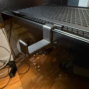 Op de afbeelding: Een zwarte hoofdtelefoonhanger bevestigd aan een computerkast. De hanger is gemaakt van plastic en heeft een gebogen ontwerp om een hoofdtelefoon vast te houden. De computerkast heeft een zwart mesh-bovenkant.