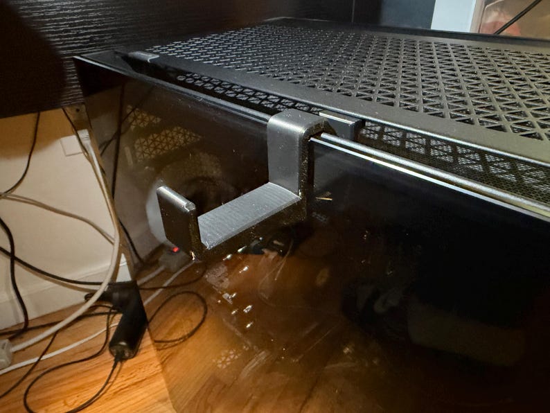 Op de afbeelding: Een zwarte hoofdtelefoonhanger bevestigd aan een computerkast. De hanger is gemaakt van plastic en heeft een gebogen ontwerp om een hoofdtelefoon vast te houden. De computerkast heeft een zwart mesh-bovenkant.