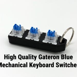 K&ouml;nnte beinhalten: Ein schwarzer, rechteckiger Schl&uuml;sselanh&auml;nger mit drei Gateron Blue mechanischen Tastaturschaltern. Die Schalter haben blaue Stiele und transparente Geh&auml;use. Ein silberner Metallring ist angebracht. Der Text lautet "High Quality Gateron Blue Mechanical Keyboard Switches".