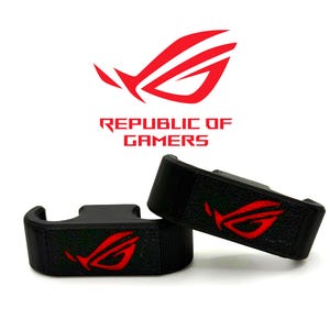 Puede incluir: Dos accesorios de juego negros con un logotipo rojo, con el texto "REPUBLIC OF GAMERS" arriba. El logotipo es un diseño rojo estilizado. Los accesorios están sobre un fondo blanco.