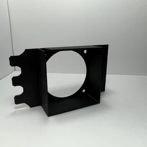 May include: A black, rectangular bracket with a large circular opening and three smaller cutouts on one side. The bracket has a box-like structure with a circular hole in the center and is made of a matte black material.