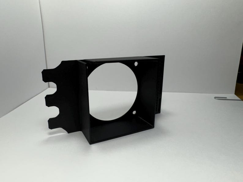 May include: A black, rectangular bracket with a large circular opening and three smaller cutouts on one side. The bracket has a box-like structure with a circular hole in the center and is made of a matte black material.