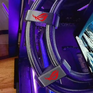 May include: A close-up of a computer's internal components, featuring a liquid cooling system with grey and black braided tubing. The system includes grey brackets with a red logo. The background is a mix of black and purple.