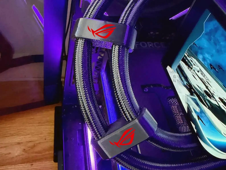 May include: A close-up of a computer's internal components, featuring a liquid cooling system with grey and black braided tubing. The system includes grey brackets with a red logo. The background is a mix of black and purple.
