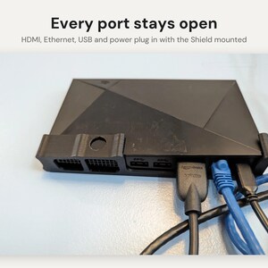 May include: A black plastic mounting bracket holding a media streaming device. The text above reads: Every port stays open. HDMI, Ethernet, USB and power plug in with the Shield mounted. The device has multiple cables connected to its rear ports.