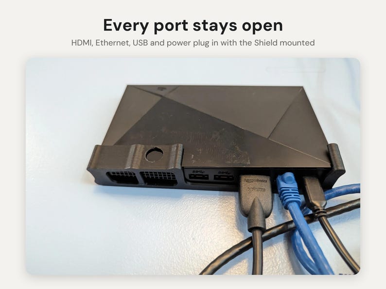 May include: A black plastic mounting bracket holding a media streaming device. The text above reads: Every port stays open. HDMI, Ethernet, USB and power plug in with the Shield mounted. The device has multiple cables connected to its rear ports.