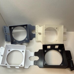 May include: Four plastic fan shrouds in different colours: grey, off-white, white, and black. Each shroud features a square frame with a circular aperture and mounting tabs. The shrouds are designed to house a fan.