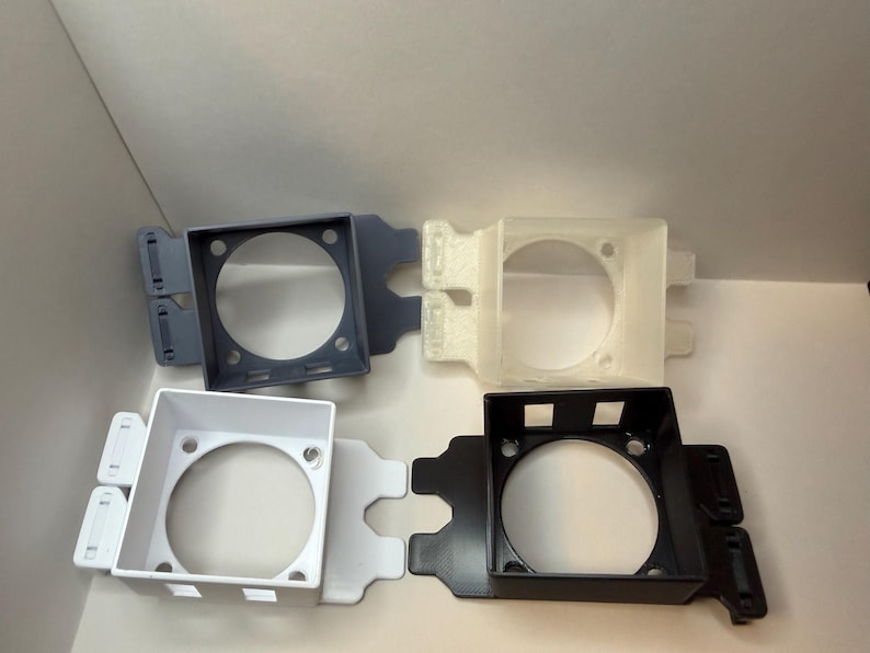 May include: Four plastic fan shrouds in different colours: grey, off-white, white, and black. Each shroud features a square frame with a circular aperture and mounting tabs. The shrouds are designed to house a fan.