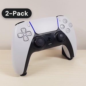 May include: A white and black wireless video game controller featuring a central touchpad, two analog sticks, a directional pad, and four action buttons. A black badge in the upper left corner displays the white text 2-Pack.