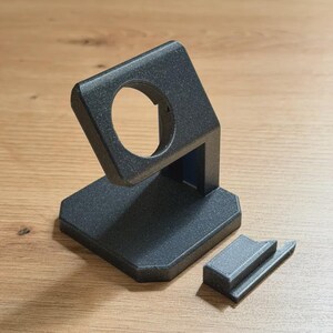 May include: A dark grey, 3D-printed watch stand with a circular opening for the watch face and a separate charging dock. The stand has a textured surface and is designed to hold a smartwatch. The base is hexagonal.