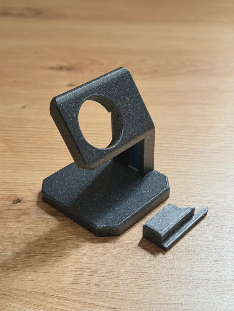 May include: A dark grey, 3D-printed watch stand with a circular opening for the watch face and a separate charging dock. The stand has a textured surface and is designed to hold a smartwatch. The base is hexagonal.