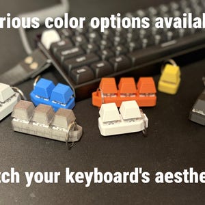 K&ouml;nnte beinhalten: Schl&uuml;sselanh&auml;nger in verschiedenen Farben, darunter Wei&szlig;, Blau, Orange, Gelb und Schwarz, in Form von Tastaturtasten. Die Schl&uuml;sselanh&auml;nger sind auf einer dunklen Oberfl&auml;che mit einer Tastatur im Hintergrund angeordnet. Der Text lautet "Various color options available" und "Match your keyboard's aesthetic!"