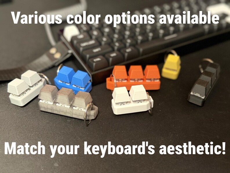 K&ouml;nnte beinhalten: Schl&uuml;sselanh&auml;nger in verschiedenen Farben, darunter Wei&szlig;, Blau, Orange, Gelb und Schwarz, in Form von Tastaturtasten. Die Schl&uuml;sselanh&auml;nger sind auf einer dunklen Oberfl&auml;che mit einer Tastatur im Hintergrund angeordnet. Der Text lautet "Various color options available" und "Match your keyboard's aesthetic!"