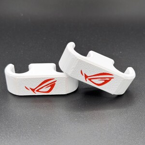 May include: Two white, 3D-printed brackets with a red logo. The brackets have a curved design and are placed on a dark surface. The red logo is a stylized eye or wing shape.