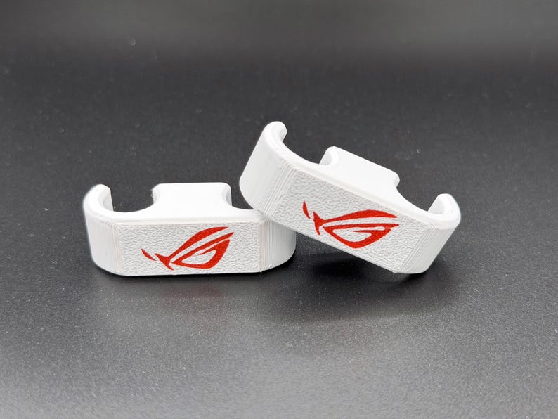 May include: Two white, 3D-printed brackets with a red logo. The brackets have a curved design and are placed on a dark surface. The red logo is a stylized eye or wing shape.