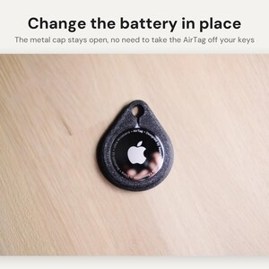 May include: A black textured protective holder for an Apple AirTag with a teardrop shape and a circular cutout at the top. The text Change the battery in place and The metal cap stays open, no need to take the AirTag off your keys is visible.