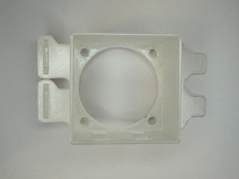 May include: A translucent, square plastic component with a central circular aperture and four small holes. It has two rectangular tabs on one side and two on the opposite. Likely a mounting bracket or housing.